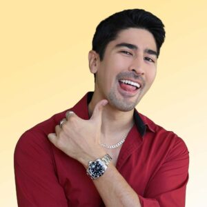 Latino Bong Juan, playfully exhibiting his iconic chain, rings, and luxury watch, smiles for the camera with tongue out.